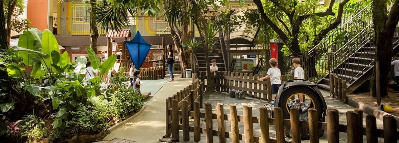 Pátio arborizado da escola com parquinho, cercas de madeira e crianças brincando