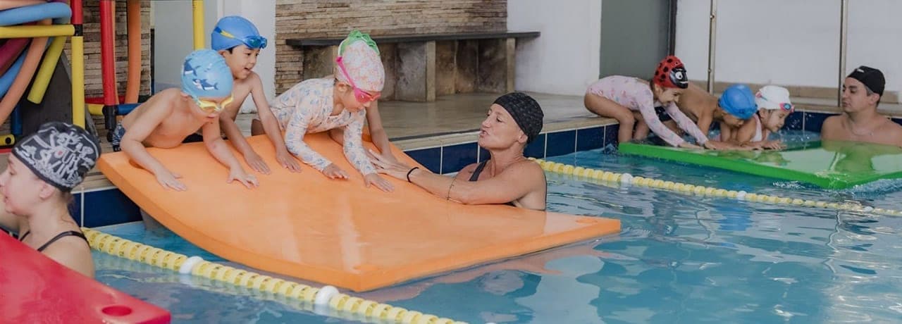 Crianças com touca e óculos de natação em aula na piscina com professores e tapetes flutuantes