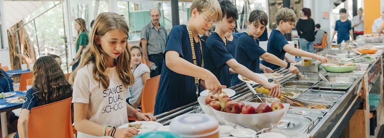 Alunos se servindo no bufê do restaurante da escola, com tigela de maçãs em primeiro plano