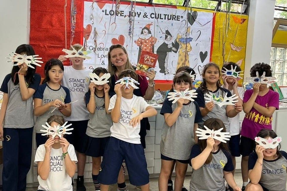 Alunos com máscaras de papel e uma professora com livro de Carnaval diante do painel "Love, Joy & Culture"