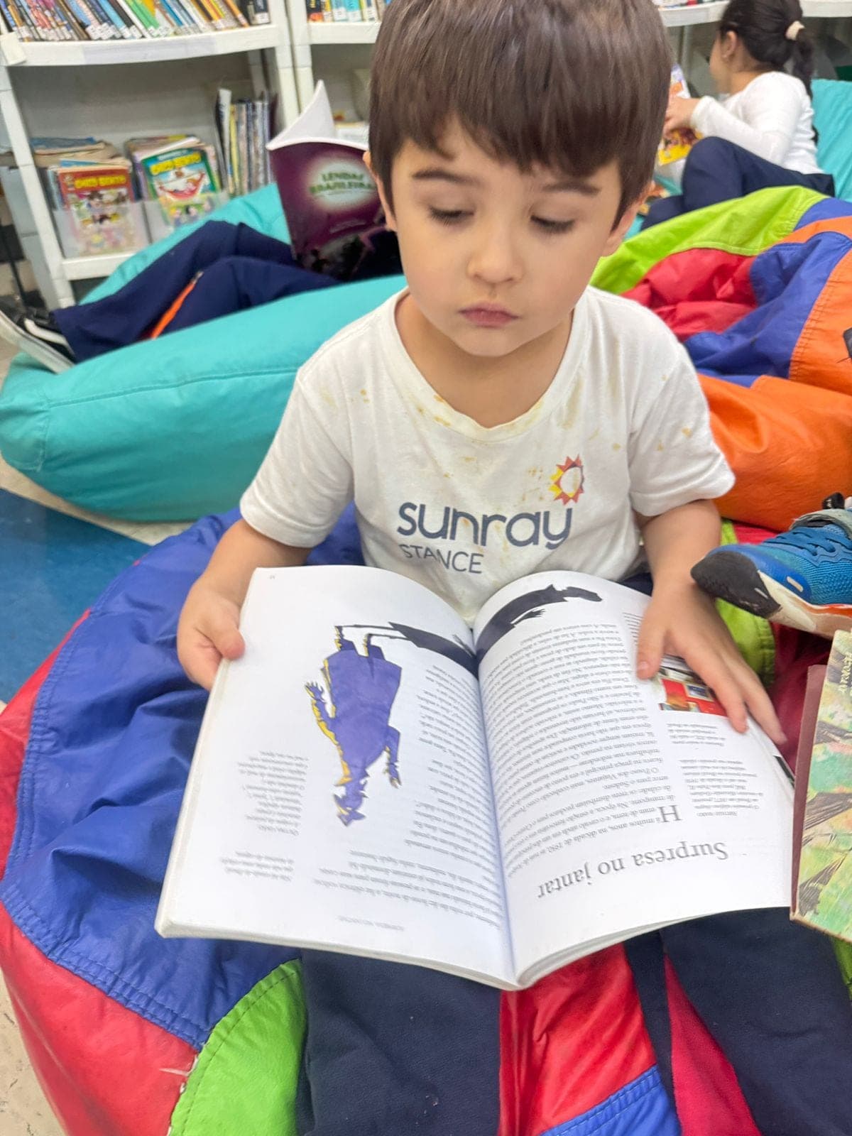 Menino sentado em um pufe lê um livro ilustrado com personagens do folclore