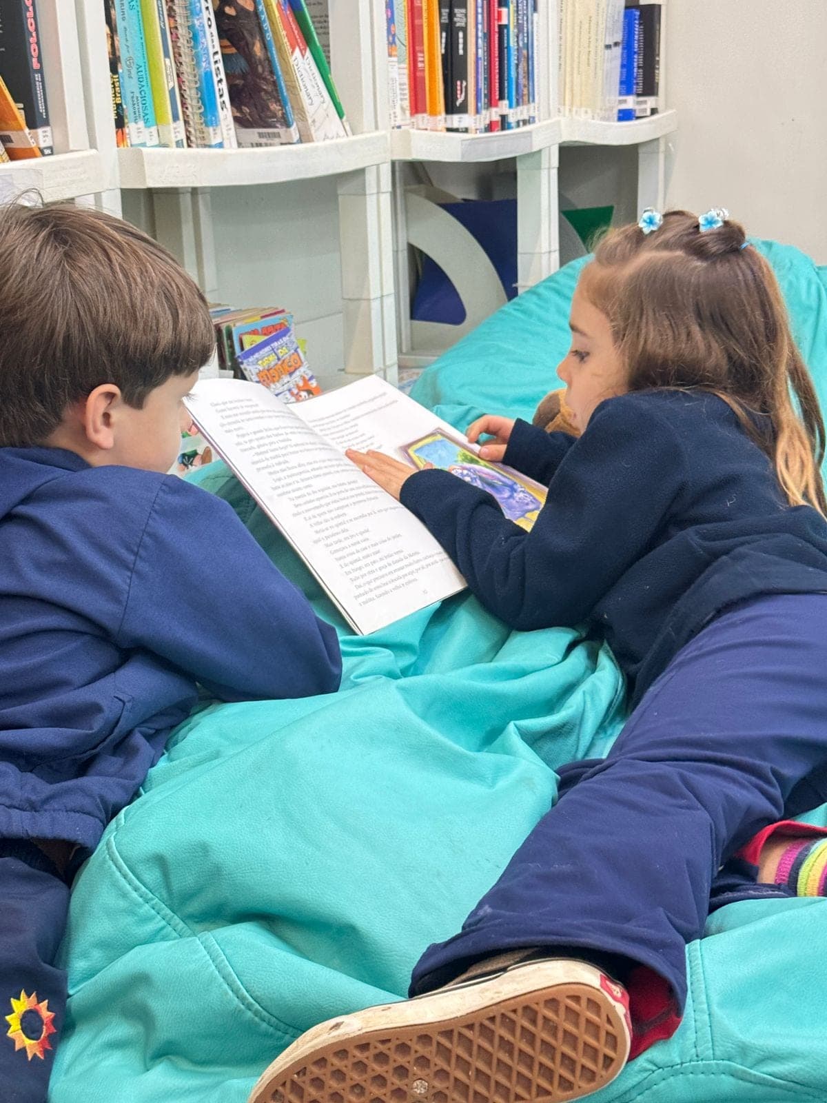 Duas crianças deitadas em pufes leem livros na biblioteca