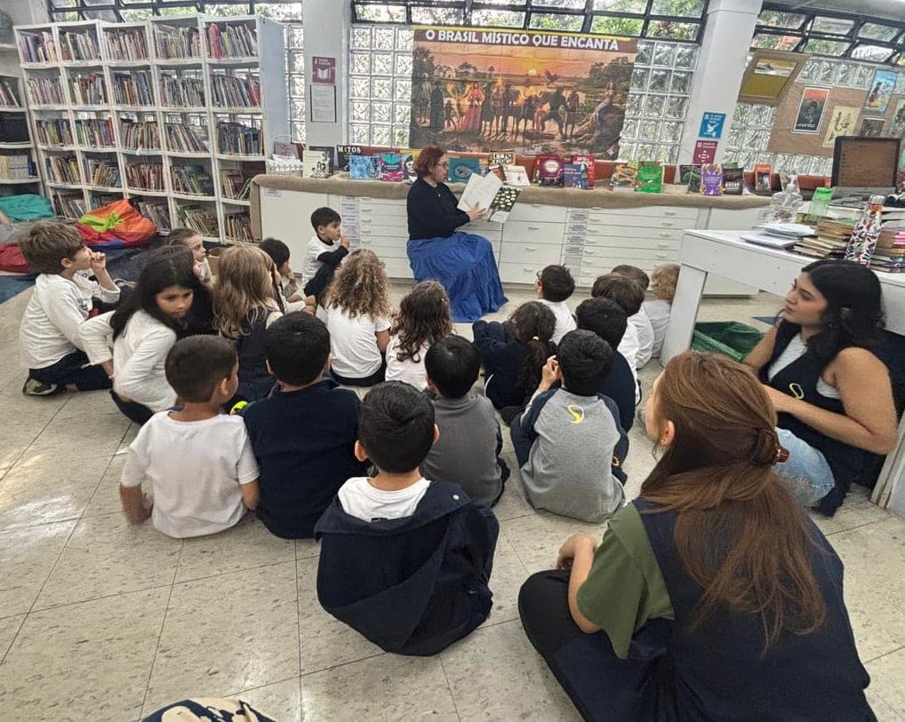 Crianças sentadas no chão da biblioteca ouvem a leitura da história, diante do painel da exposição