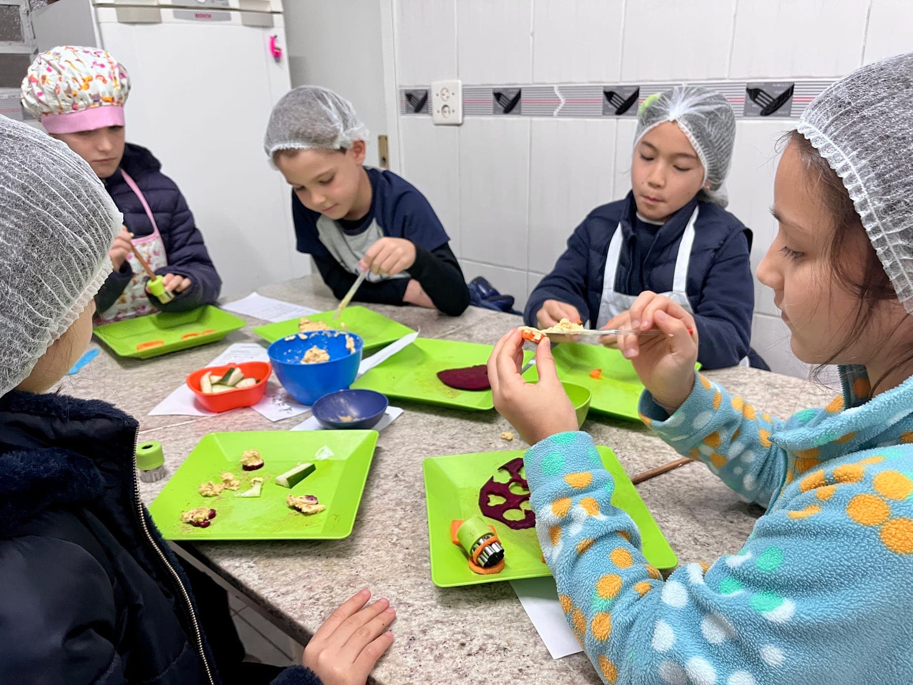 Alunos à mesa preparam fatias de cenoura, beterraba e pepino sobre bandejas verdes