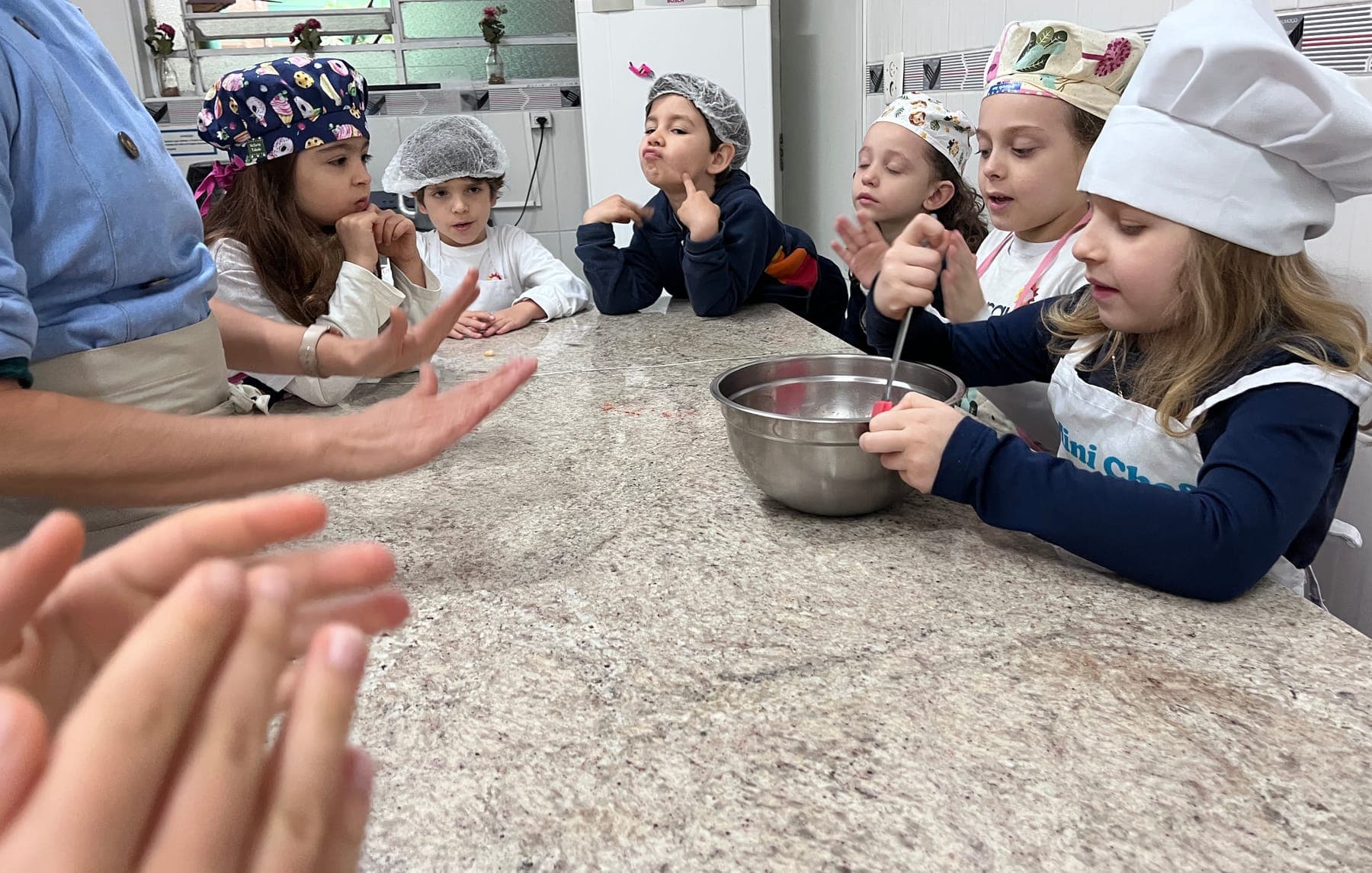 Turma do K5 de touca reunida na bancada com tigelas, enquanto a professora explica a receita