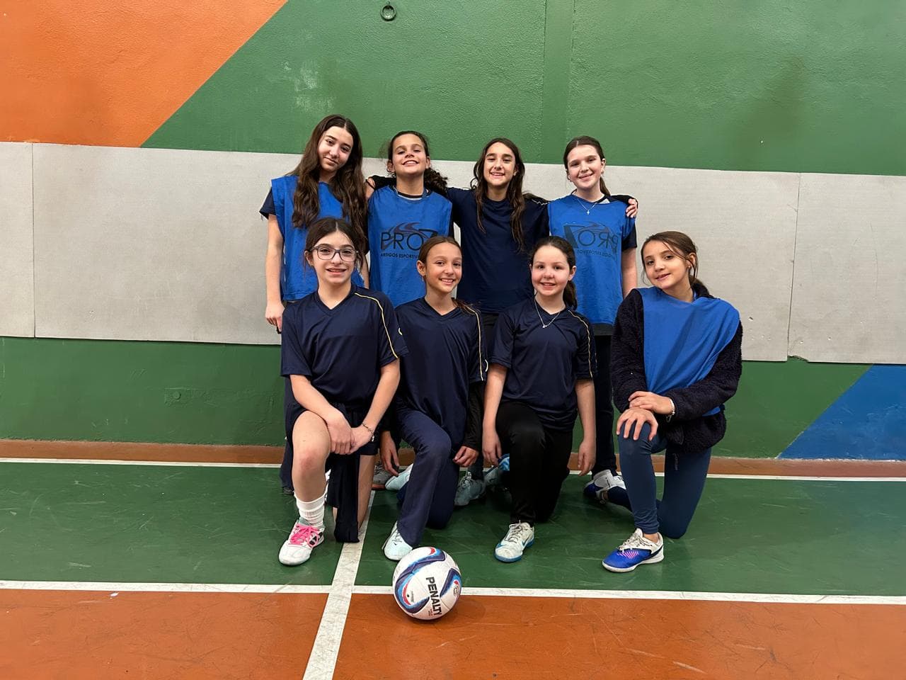 Equipe de alunas com coletes azuis posando com a bola na quadra de futsal