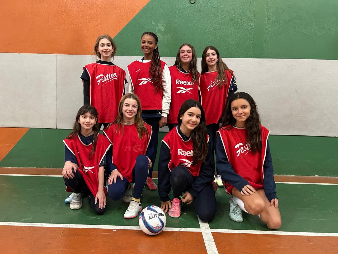Equipe de alunas com coletes vermelhos posando com a bola na quadra de futsal