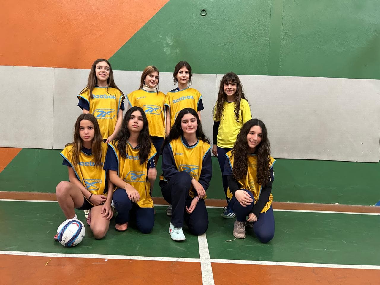 Equipe de alunas com coletes amarelos posando com a bola na quadra de futsal