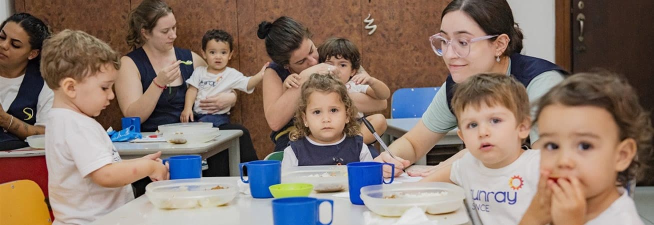 Crianças pequenas fazendo a refeição à mesa com a ajuda das educadoras