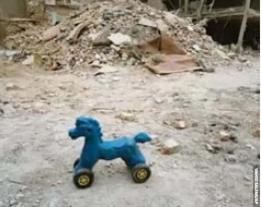 Cavalinho de brinquedo azul sobre o chão de terra diante de escombros