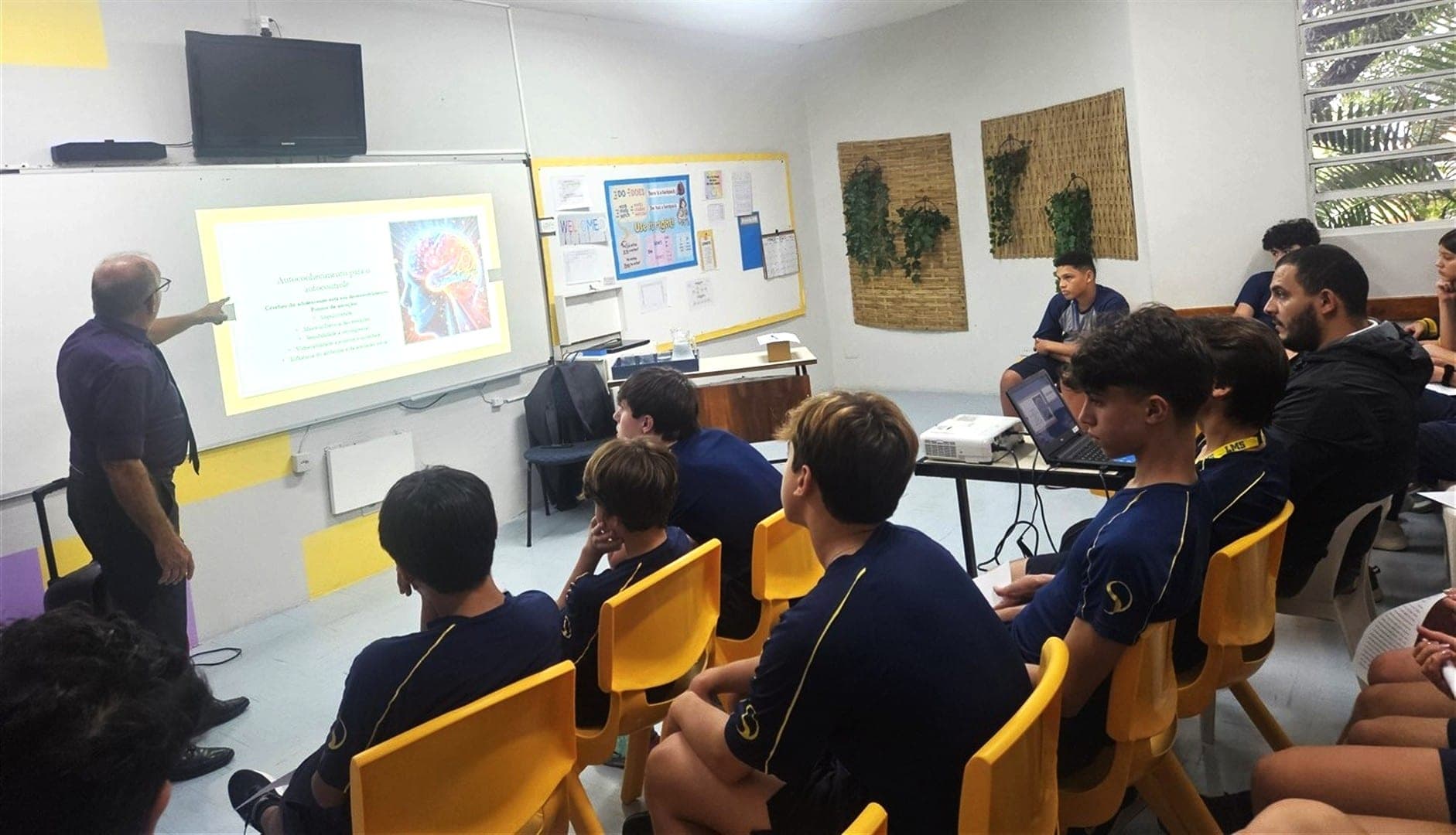 Palestrante apresenta slides a alunos adolescentes sentados em cadeiras amarelas na sala de aula