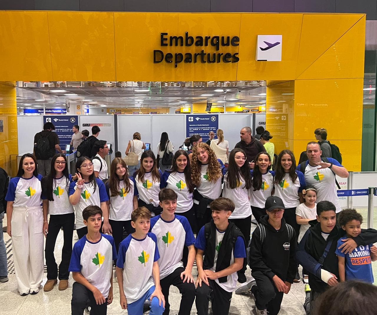 Grupo de alunos e acompanhantes com camisetas de folha de bordo verde e amarela no portão de embarque do aeroporto