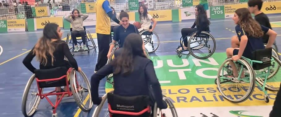 Alunas jogando basquete em cadeiras de rodas em quadra com faixa Juntos pelo esporte paralímpico