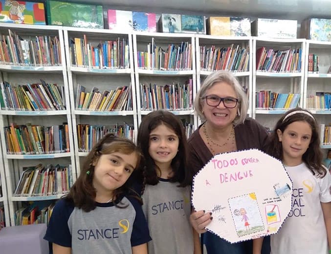 Três alunas e uma educadora na biblioteca seguram cartaz desenhado com a frase "Todos contra a dengue"