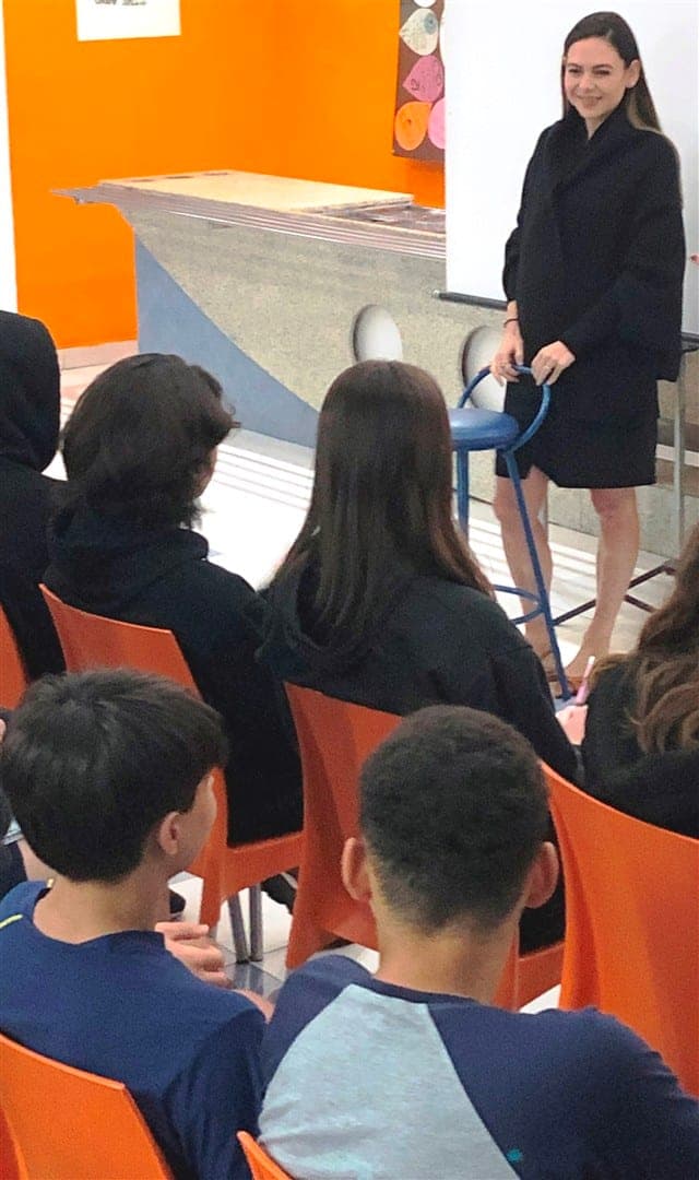 Isadora Cohen, de pé ao lado de um banco, conversa com alunos sentados em cadeiras laranja