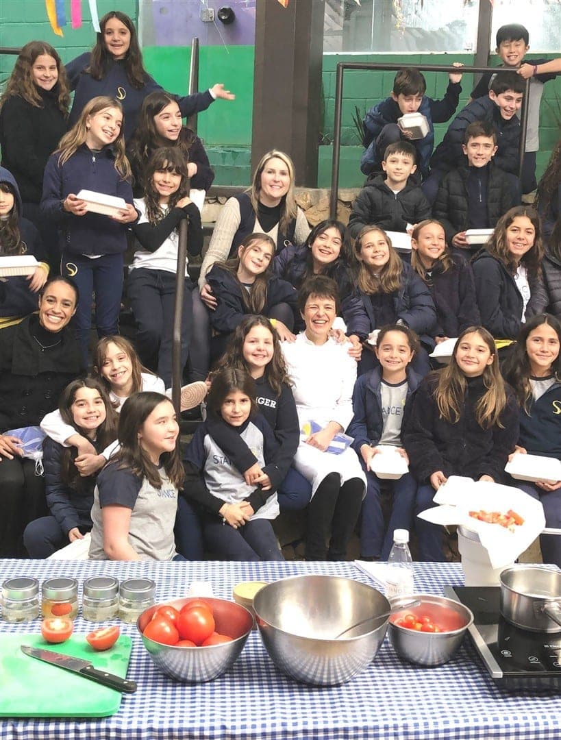 Chef Vivi Gonçalves posa com a turma do 5º ano atrás de uma mesa com tomates e utensílios de cozinha