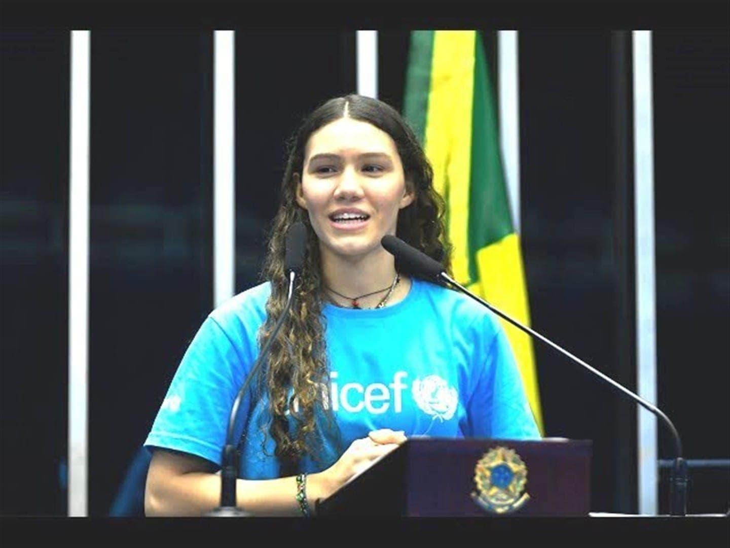 Catarina Lorenzo, com camiseta do UNICEF, discursa em uma tribuna diante da bandeira do Brasil