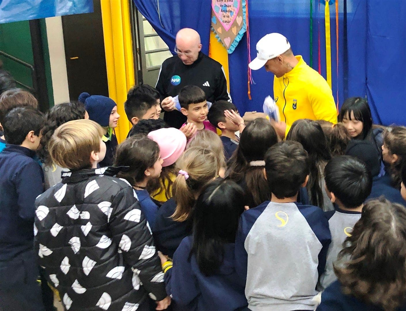 Kau Claro, de agasalho amarelo, autografa papéis cercado por alunos da Stance