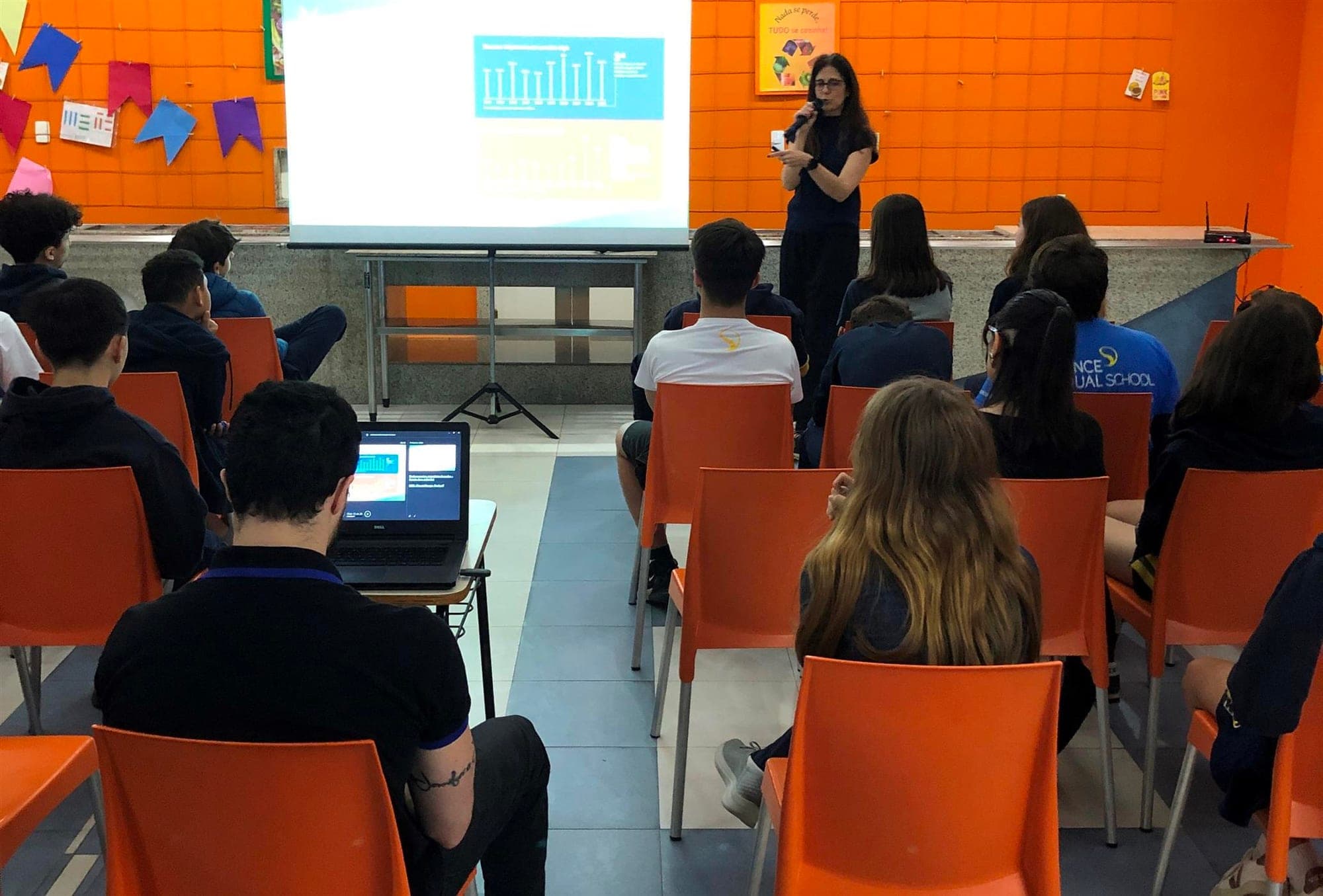 Dra. Maria Christina Gueorguiev fala ao microfone para alunos do 8º e 9º ano diante de um telão
