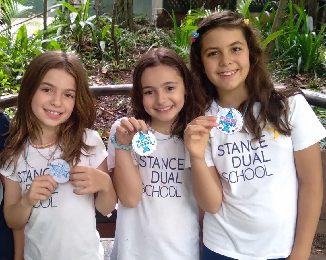 Três alunas com camiseta da Stance Dual School mostram medalhas de papel com a frase "Mares limpos"