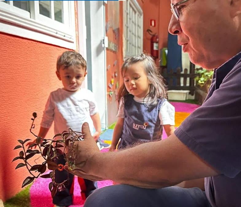 Educador mostra uma muda de planta a duas crianças pequenas no espaço da Educação Infantil