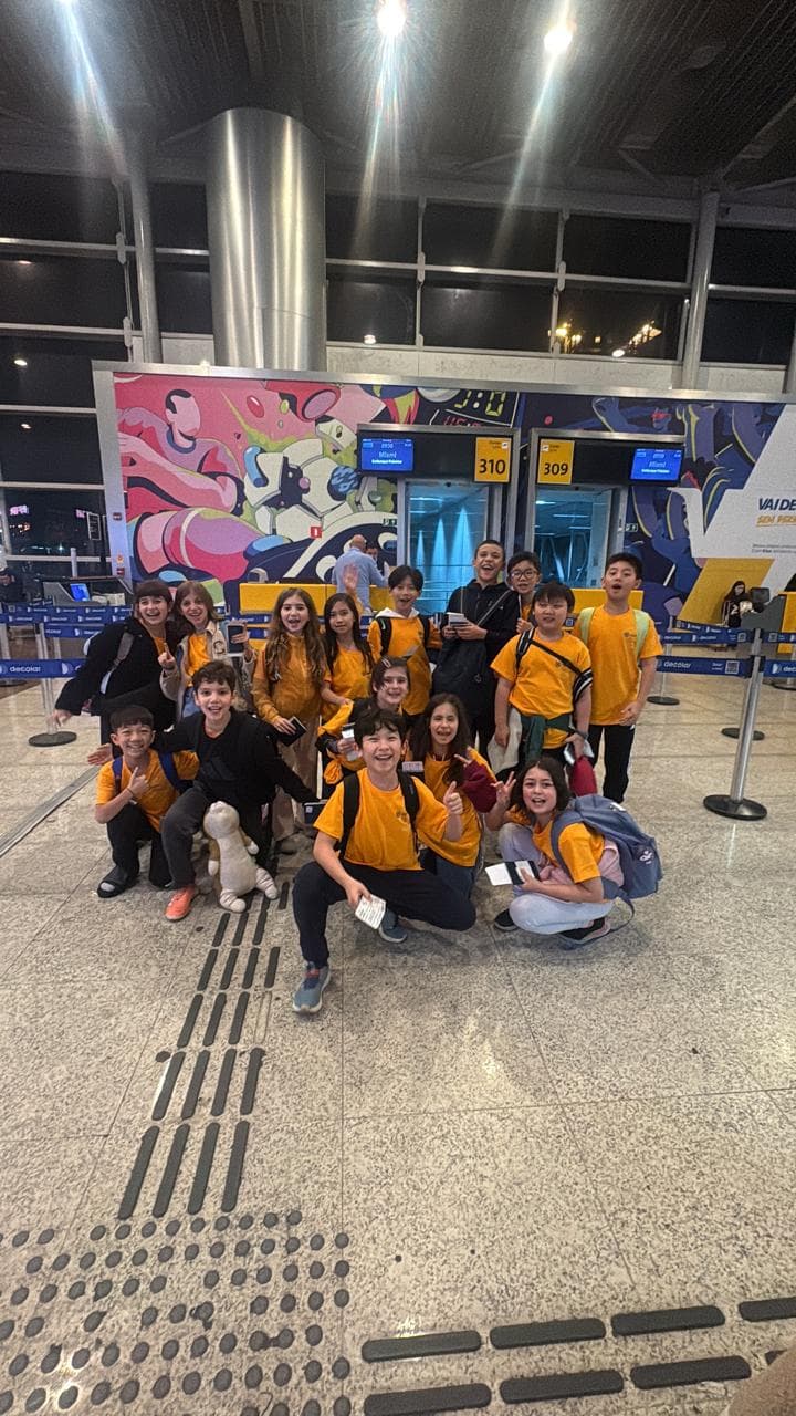 Grupo de alunos com camisetas amarelas e mochilas posa animado no saguão do aeroporto, diante do portão de embarque