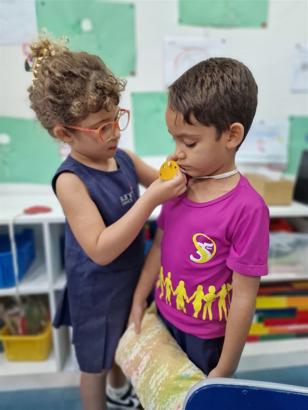Menina de óculos ajusta um colar de brinquedo amarelo no pescoço de um colega na sala