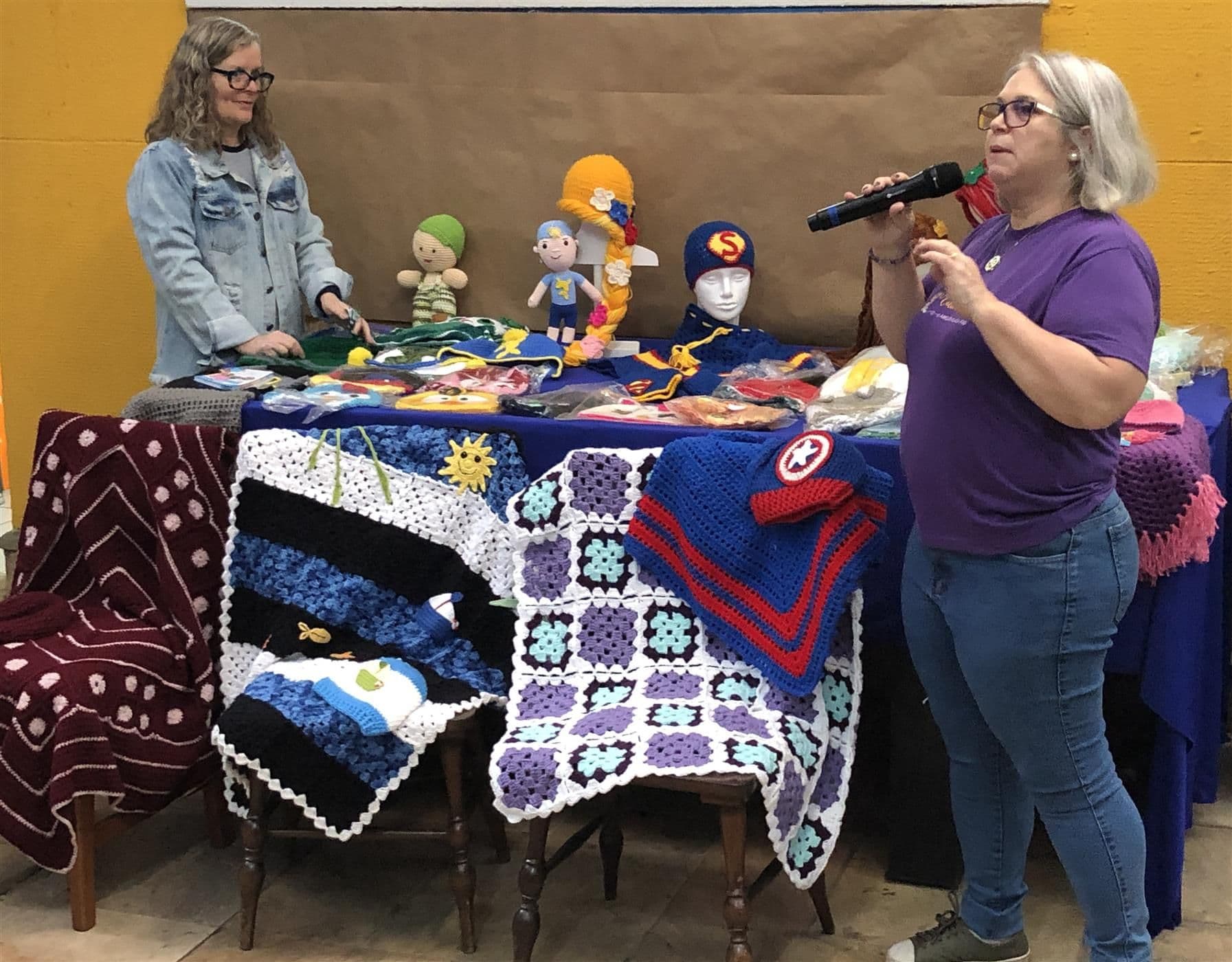 Adriana apresenta mantas, gorros e bonecos de crochê do Projeto Entrelaçadas expostos sobre mesa e cadeiras