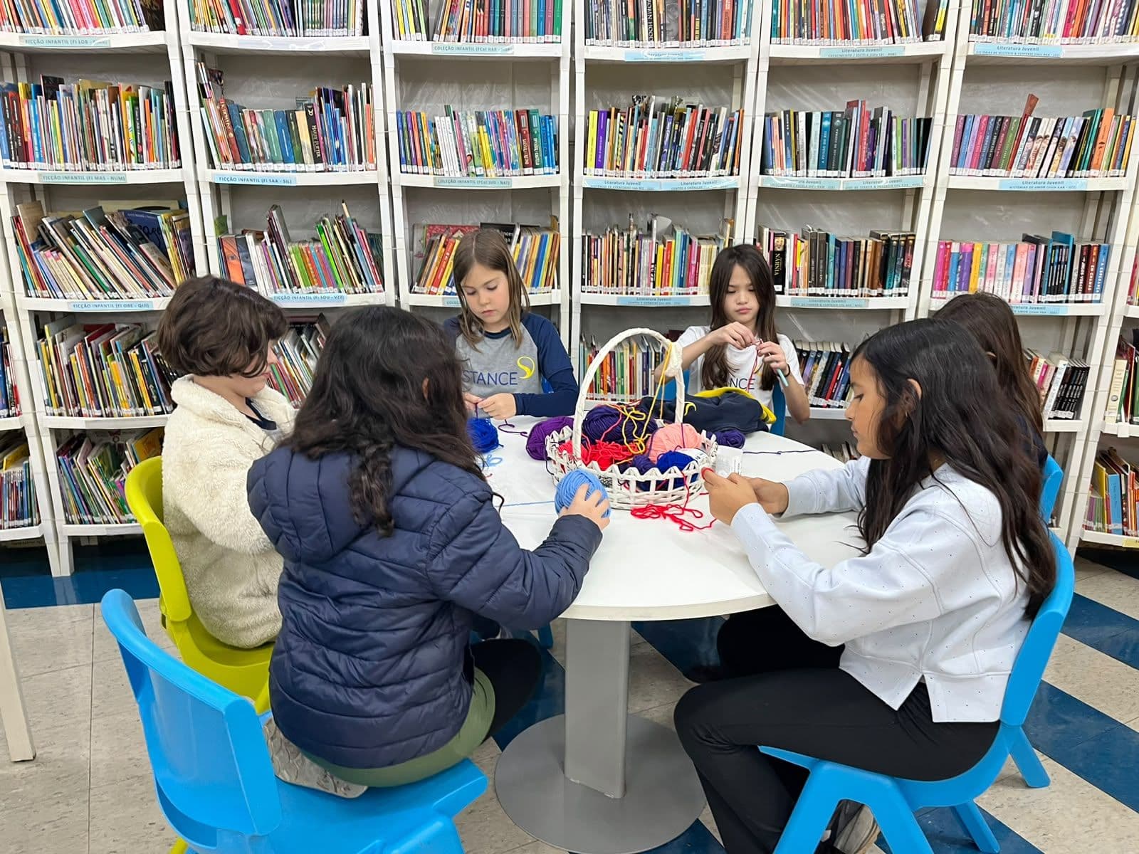 Grupo de alunos tricota com lã ao redor de uma mesa redonda, entre estantes de livros da biblioteca