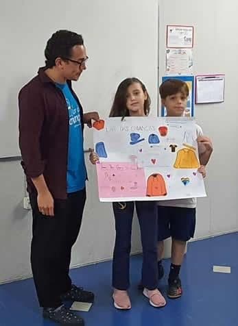 Alunos do 5º B mostram cartaz ilustrado com roupas de frio ao lado de representante do Lar das Crianças