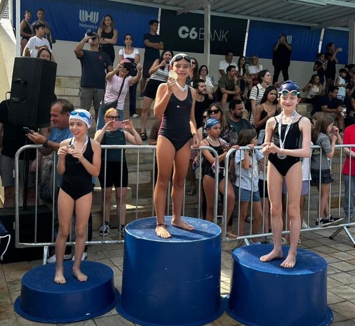 Nadadoras com medalhas no pódio da natação nas Olimpíadas da Hebraica, diante da arquibancada