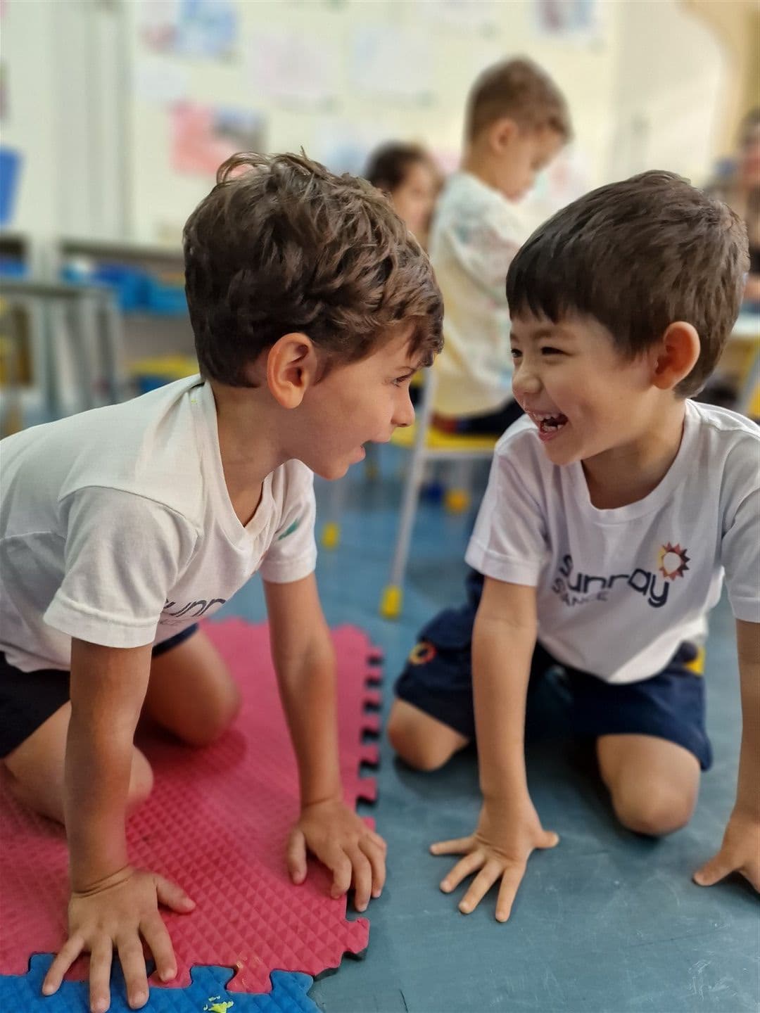 Dois meninos ajoelhados no tapete da sala de aula, frente a frente, rindo juntos
