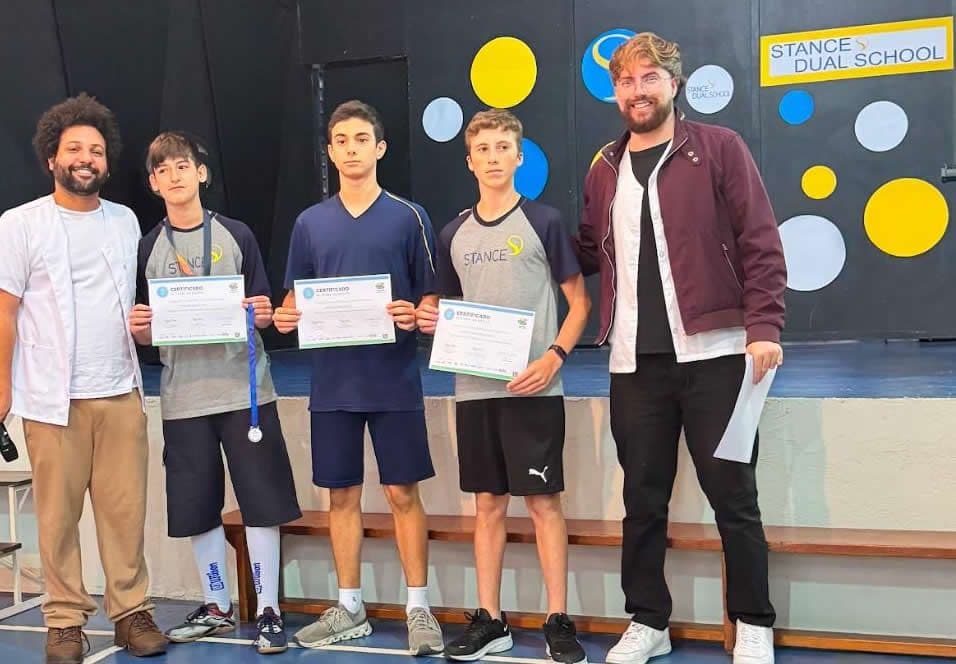 Três alunos exibem certificados e medalha no palco, ao lado de dois professores