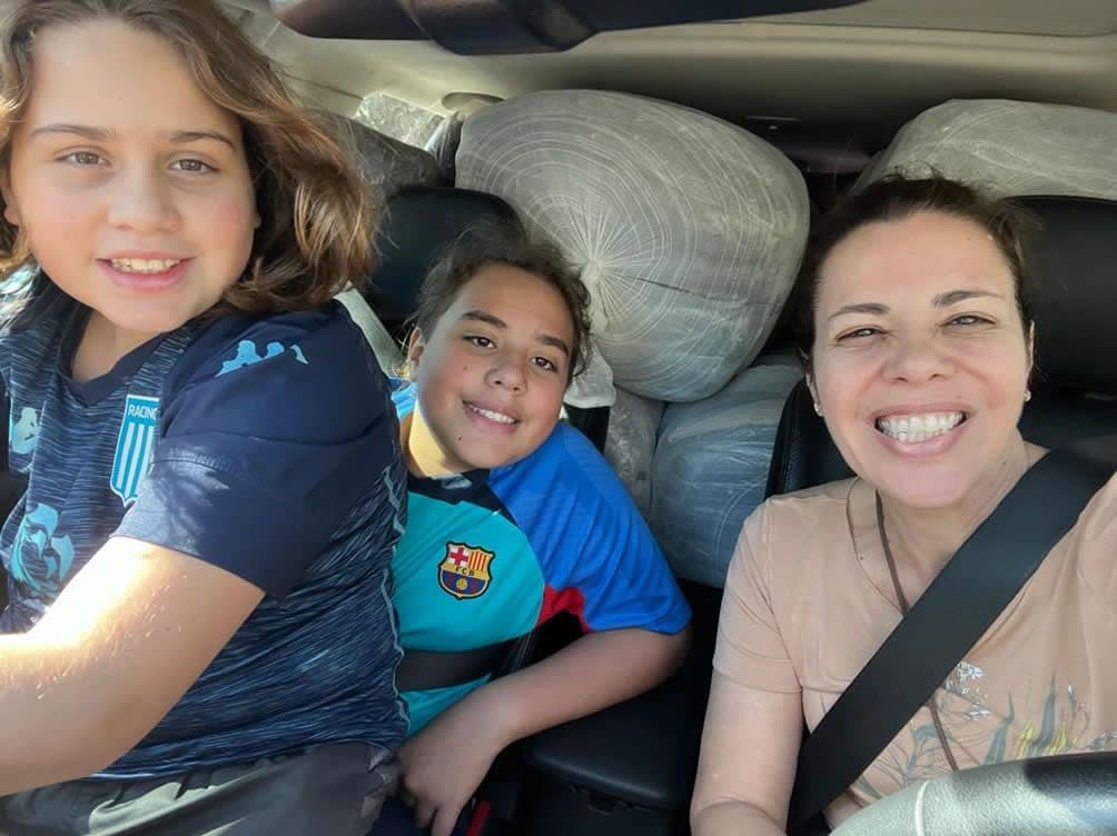 Duas crianças e uma mulher sorriem dentro do carro carregado de fardos de cobertores para doação