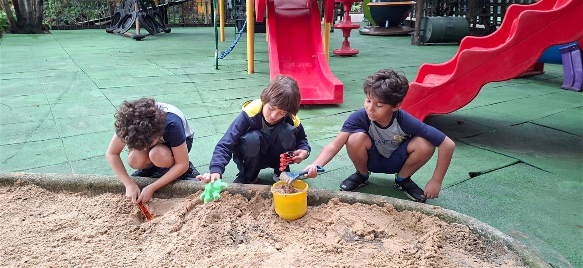 Três meninos brincam com baldes e pás no tanque de areia do parque