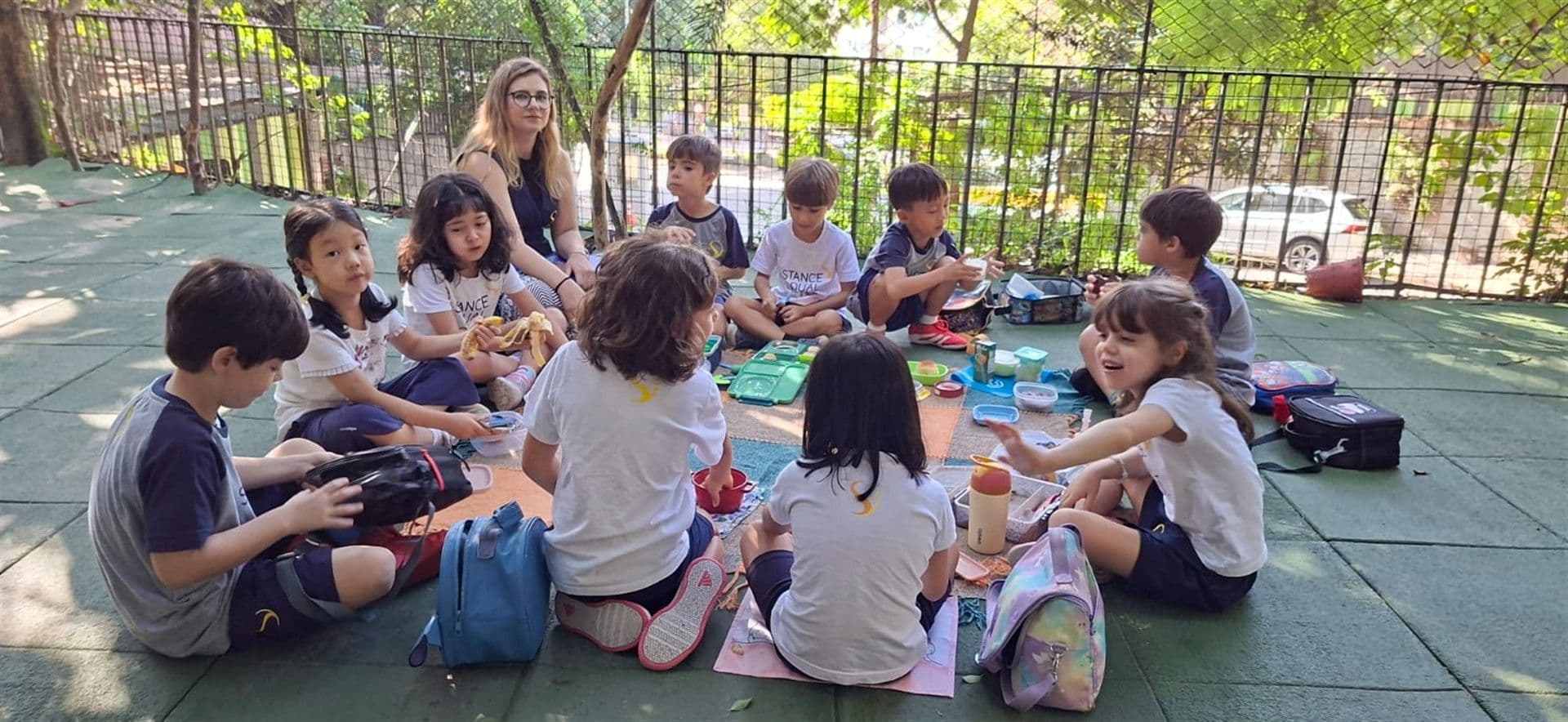 Crianças sentadas em roda no pátio fazem lanche junto com uma professora