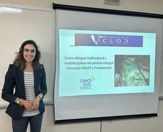 Professora Jeanny ao lado de slide do 14º Congresso ICLOC com a apresentação da Stance Dual School