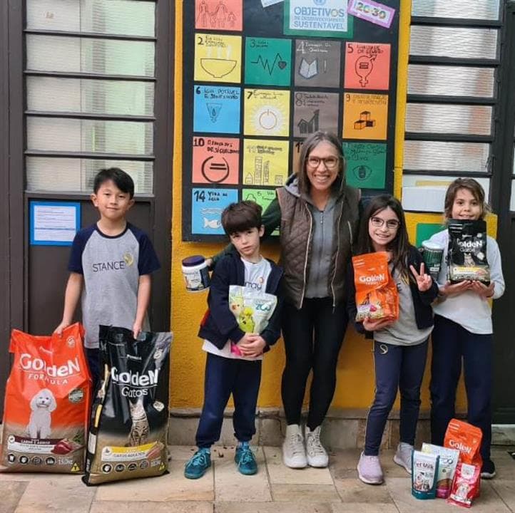 Alunos e professora com pacotes de ração doados, diante de painel dos Objetivos de Desenvolvimento Sustentável