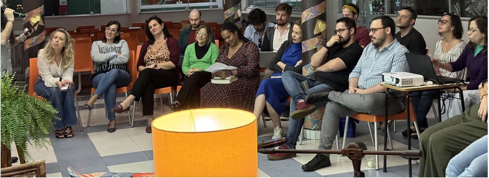 Plateia de professores sentada em cadeiras laranja acompanha as leituras do sarau