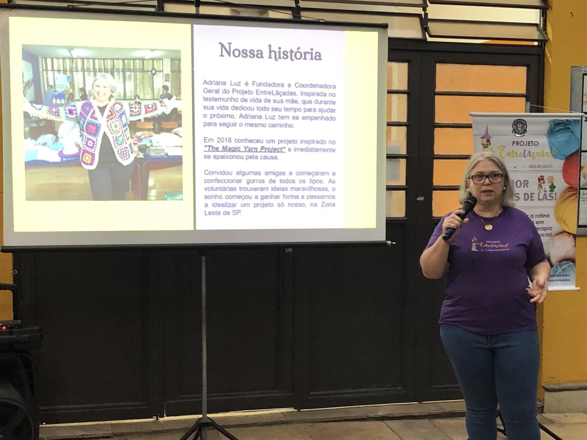 Adriana fala ao microfone ao lado do slide Nossa história, sobre a criação do Projeto Entrelaçadas