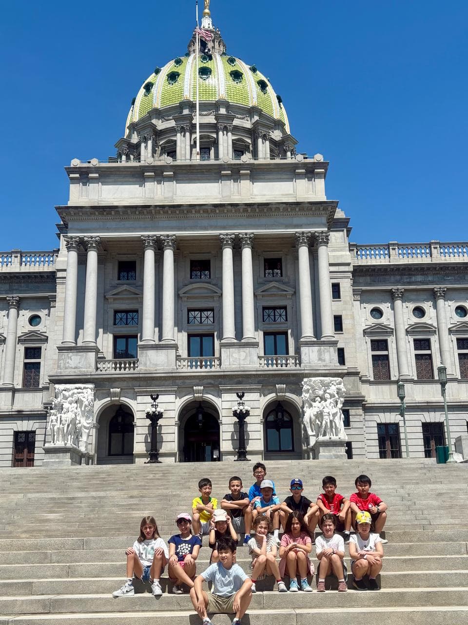 Alunos sentados na escadaria de um prédio histórico com cúpula verde, sob céu azul