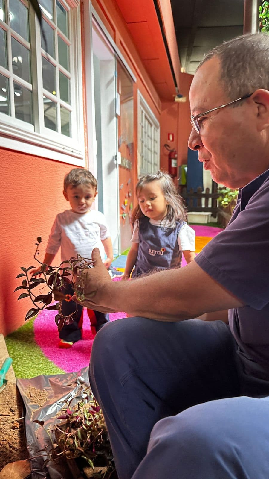 Adulto mostra uma muda de planta a duas crianças pequenas durante atividade de jardinagem