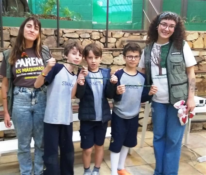 Três alunos posam com duas voluntárias do Greenpeace, uma com camiseta "Parem as máquinas"