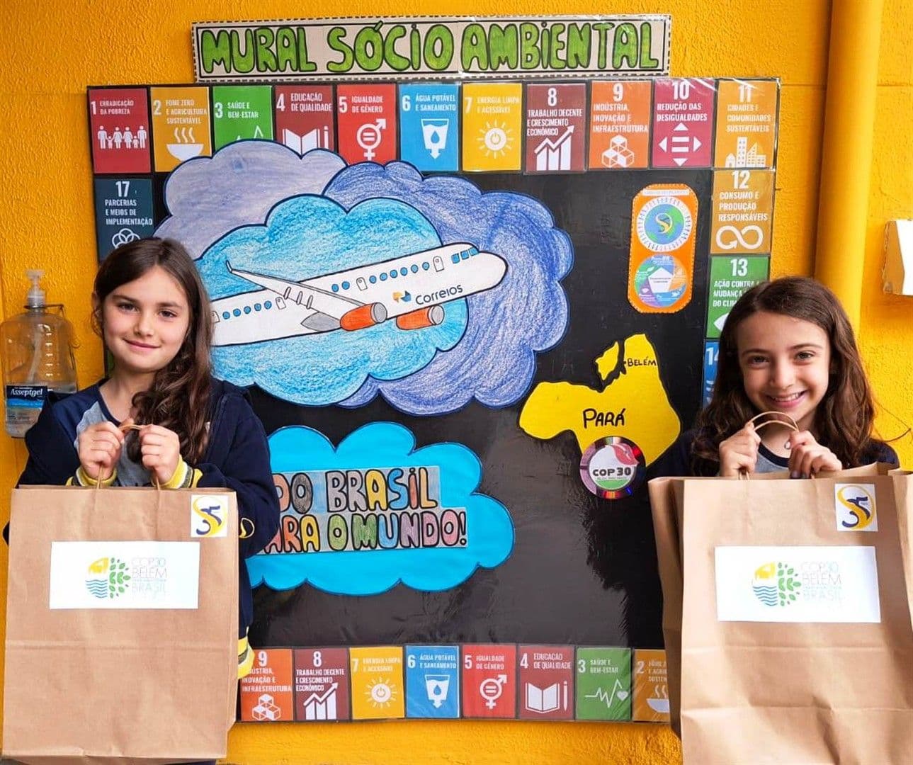 Duas alunas seguram sacolas da COP30 diante do Mural Socioambiental com os ODS e a frase Do Brasil para o mundo