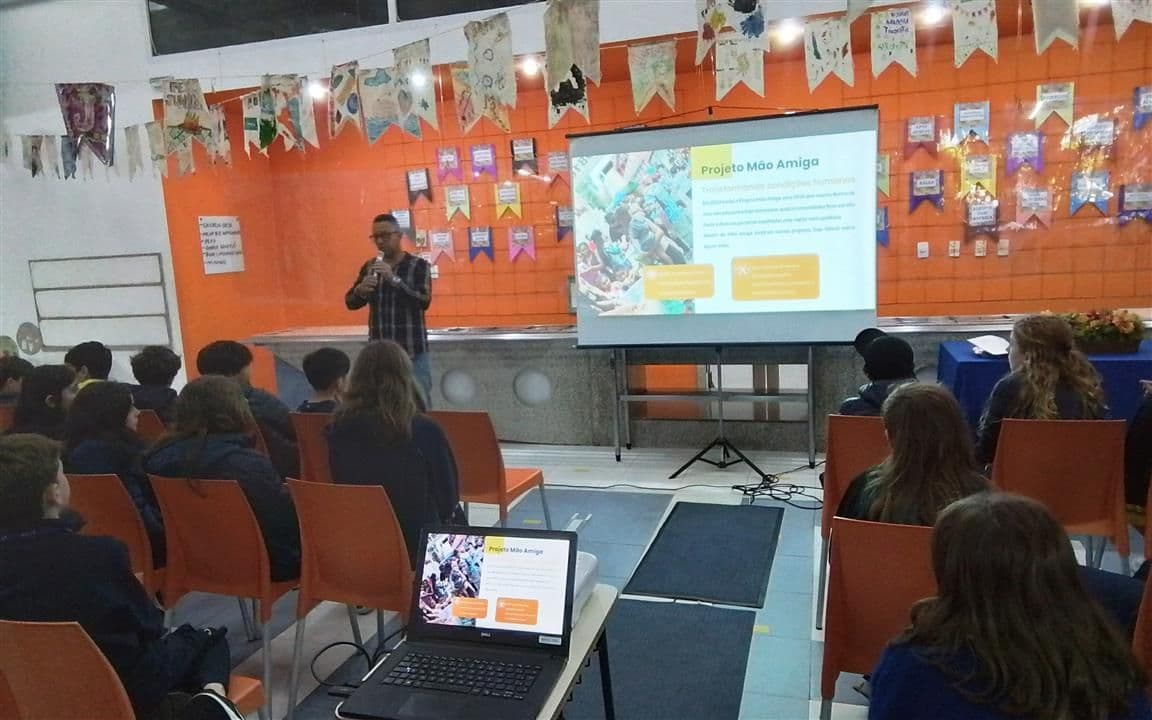 Palestrante apresenta o Projeto Mão Amiga a alunos sentados diante de um telão