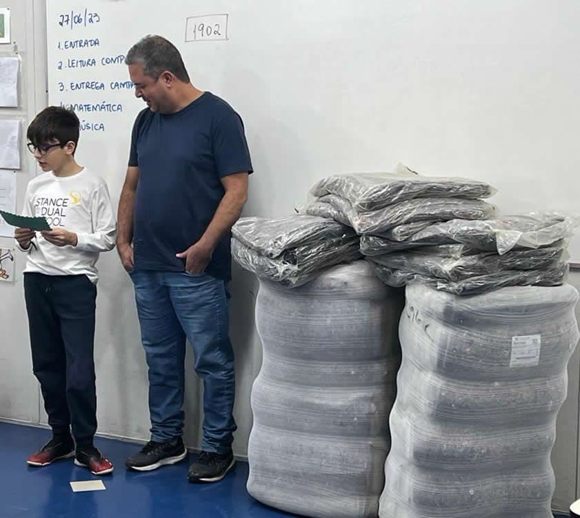 Aluno lê mensagem ao lado de representante de instituição e de fardos de cobertores doados na sala de aula