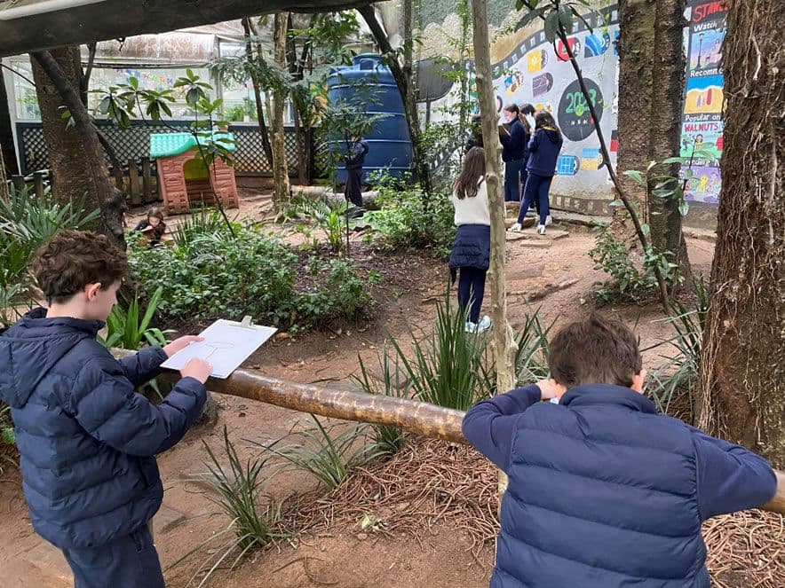 Alunos desenham em pranchetas no jardim da escola, observando árvores e plantas