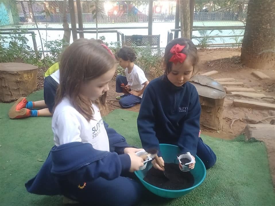 Duas meninas enchendo copinhos com terra para plantio, sentadas na grama do parque
