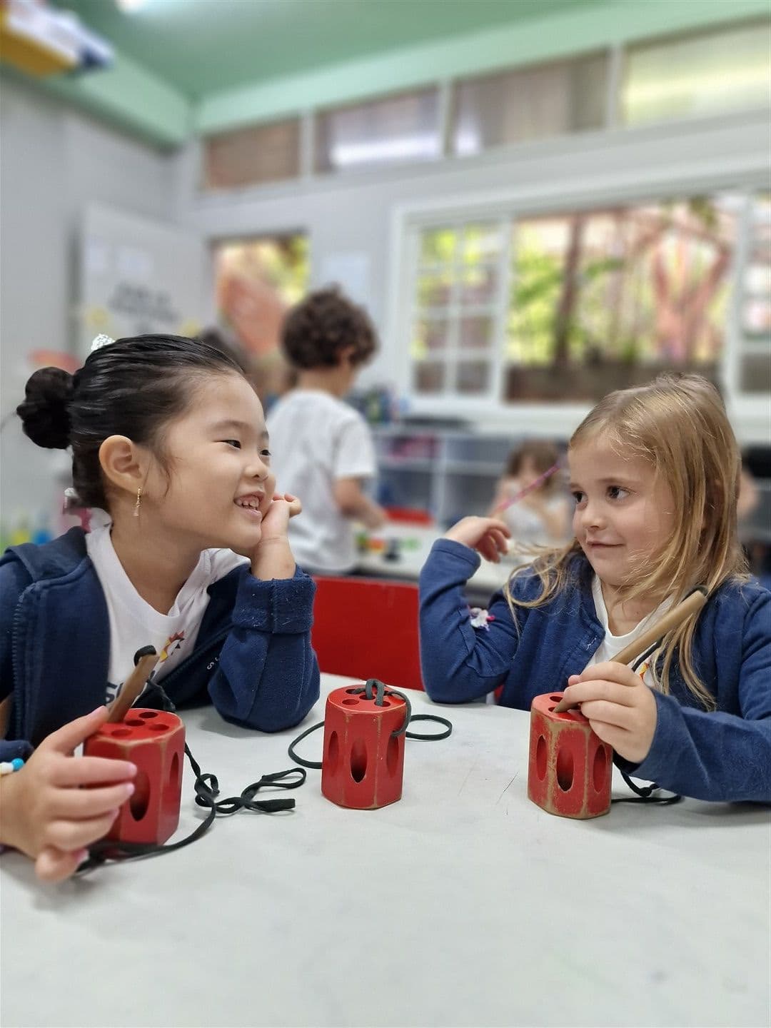 Duas meninas sorrindo uma para a outra à mesa, com blocos vermelhos de alinhavo
