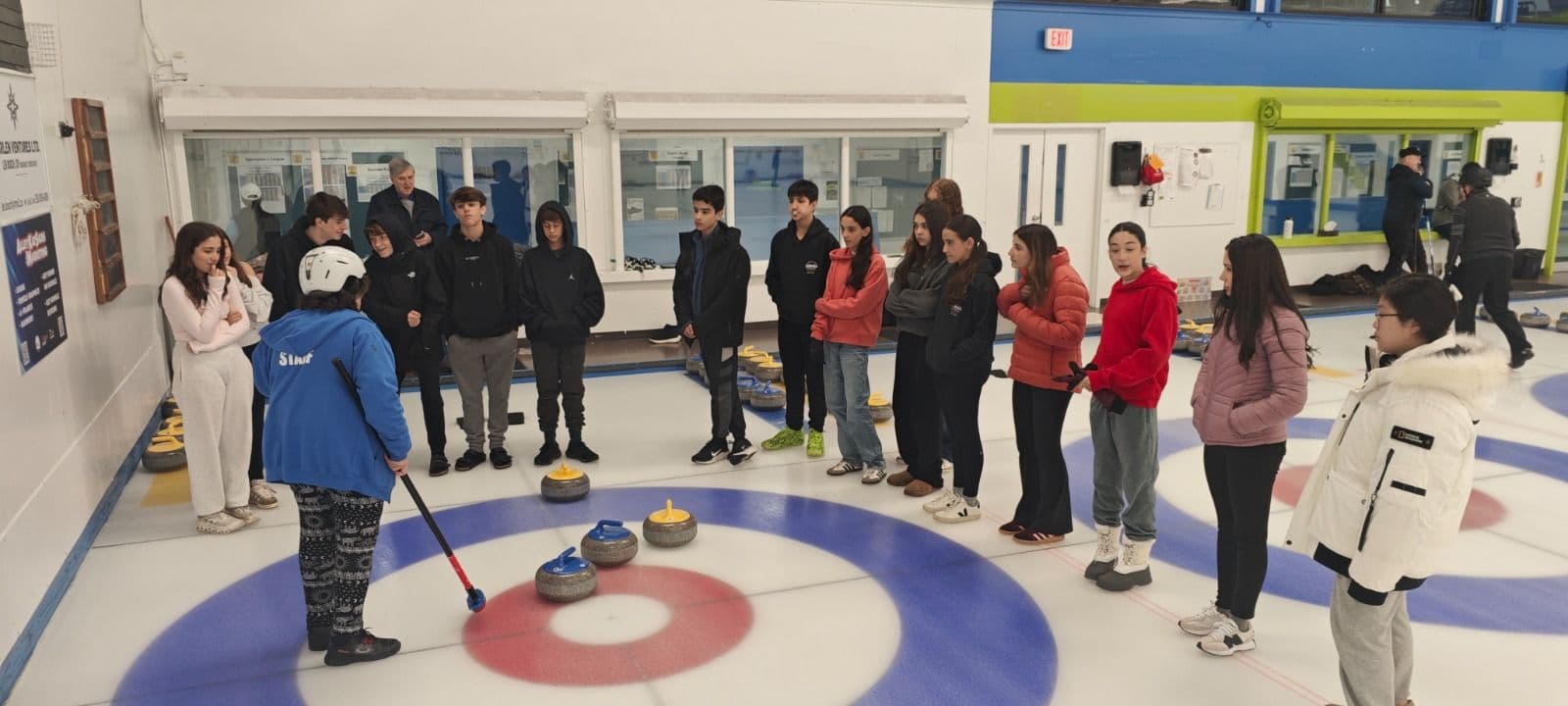 Alunos observam uma instrutora explicar o curling na pista de gelo